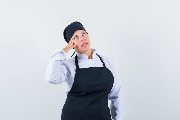 Blonde woman standing in thinking pose, leaning cheek on hand in black cook uniform and looking pensive.