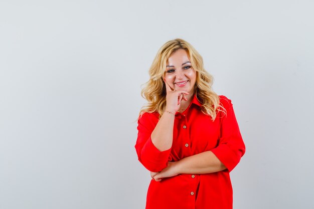 Blonde woman standing in thinking pose in red blouse and looking pensive.