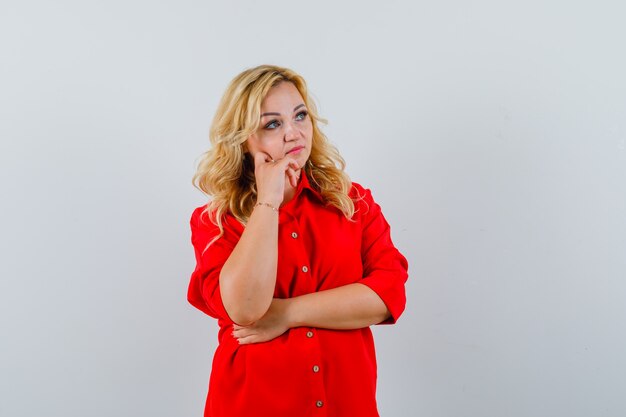 Blonde woman standing in thinking pose in red blouse and looking pensive.