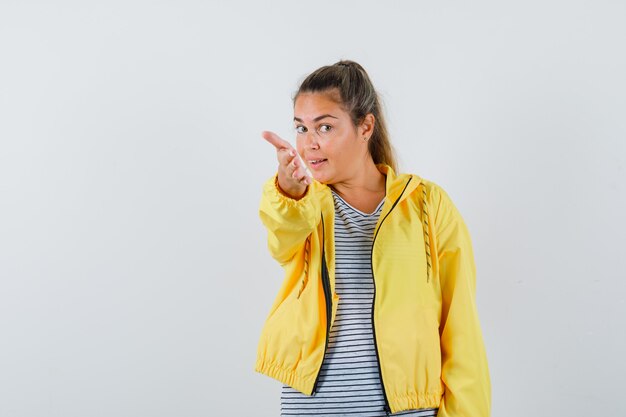 Blonde woman stretching hand toward front as inviting to come in yellow bomber jacket and striped shirt and looking pretty