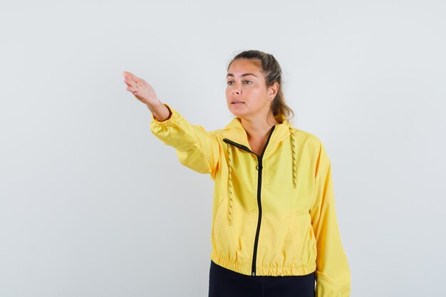 Blonde woman stretching hand toward left in yellow bomber jacket and black pants and looking serious