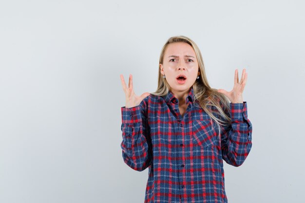 Blonde woman stretching hands and becoming nervous in checked shirt and looking shocked , front view.