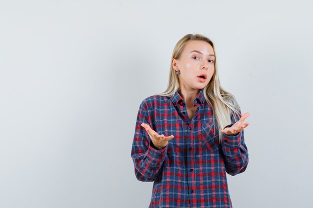 Blonde woman stretching hands while talking in checked shirt and looking curious. front view.