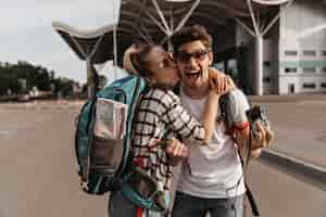 Free photo blonde woman in striped shirt kisses happy emotional man in sunglasses couple of travelers poses with retro camera and backpacks near airport