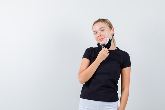 Blonde woman taking off mask, smiling in black t-shirt, white pants