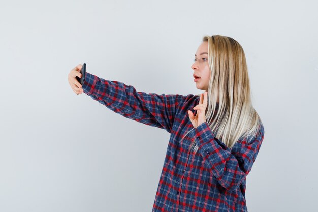 Blonde woman taking selfie and showing peace sign in checked shirt and looking attractive , front view.