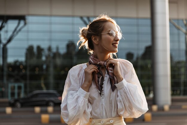 Blonde woman touches her silk brown scarf Attractive young girl in eyeglasses and white blouse smiles outside