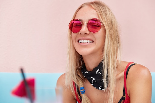 blonde woman in trendy sunglasses, wears fashionable clothing and red sunglasses, sits against pink wall on comfortable couch.