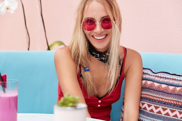 blonde woman in trendy sunglasses, wears fashionable clothing and red sunglasses, sits against pink wall on comfortable couch.