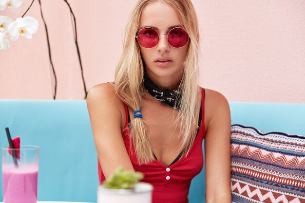 Free photo blonde woman in trendy sunglasses, wears fashionable clothing and red sunglasses, sits against pink wall on comfortable couch.