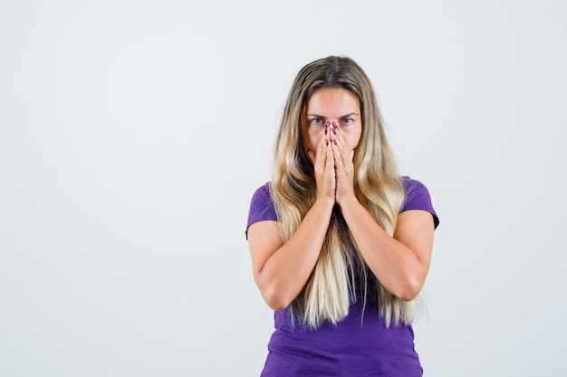 blonde woman in violet t-shirt clasping hands on face and looking hopeful , front view.