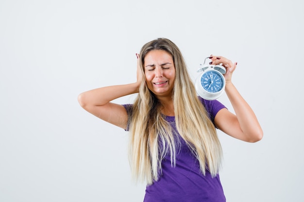 Free photo blonde woman in violet t-shirt holding alarm clock while crying and looking forgetful , front view.