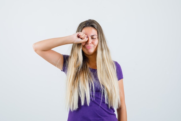 блондинка в фиолетовой футболке трет глаза, плача и обижаясь, вид спереди.