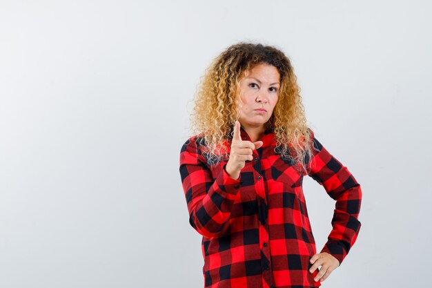 Blonde woman with curly hair in checked shirt pointing at camera and looking serious , front view.