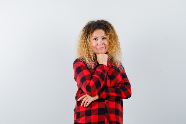 Blonde woman with curly hair in checked shirt putting hand propping up on chin and looking sensible , front view.