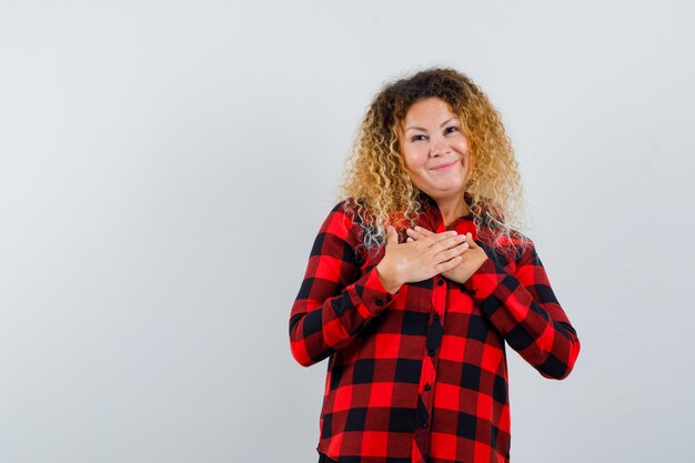 Blonde woman with curly hair keeping hands on chest in checked shirt and looking grateful. front view.