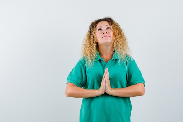 Blonde woman with curly hair showing namaste gesture in green T-shirt and looking hopeful. front view.