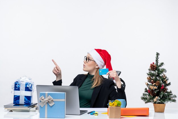 サンタクロースの帽子と眼鏡をかけて、オフィスでクリスマスプレゼントと銀行カードを保持しているテーブルに座っているブロンドの女性