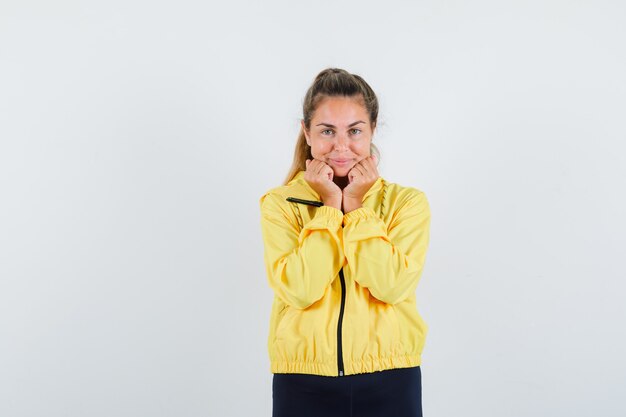 Blonde woman in yellow bomber jacket and black pants clenching fists and putting hands under chin and looking happy