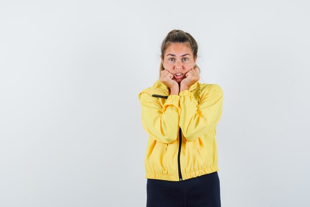 Blonde woman in yellow bomber jacket and black pants holding hands on cheeks, clenching fists and looking excited