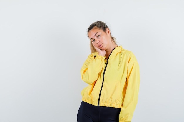 Blonde woman in yellow bomber jacket and black pants leaning cheek on palm and looking pretty