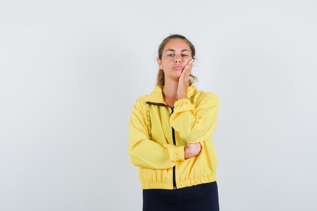 Blonde woman in yellow bomber jacket and black pants leaning cheek on palm, thinking about something and looking pensive