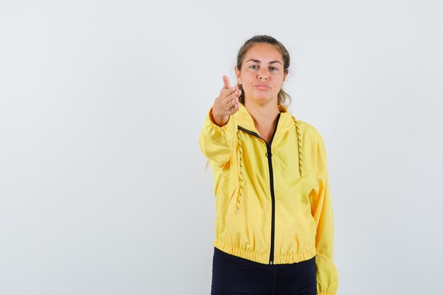 Blonde woman in yellow bomber jacket and black pants pointing at front with hand and looking serious
