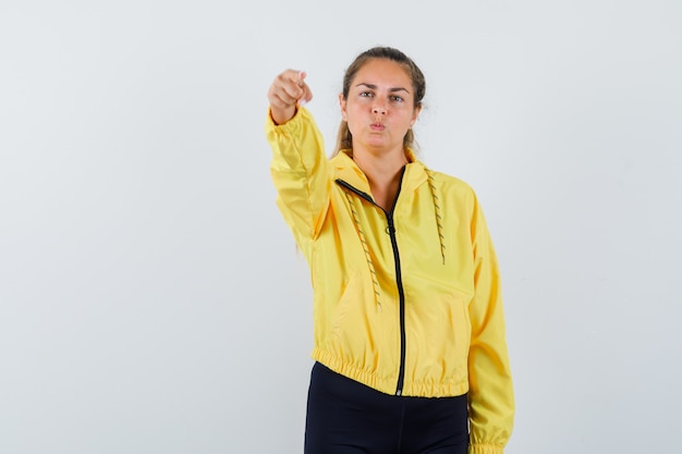 Donna bionda in bomber giallo e pantaloni neri che punta davanti con il dito indice mentre curva le labbra e sembra seria