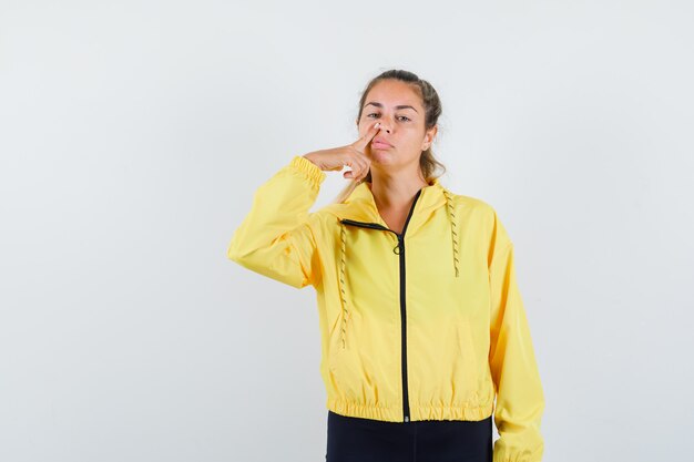 Blonde woman in yellow bomber jacket and black pants putting index finger on nose and looking pretty