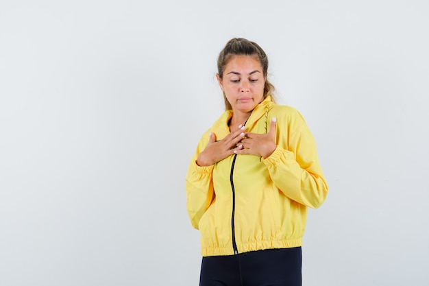 Blonde woman in yellow bomber jacket and black pants resting hands on chest and looking serious