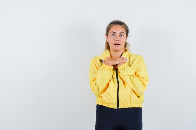 Blonde woman in yellow bomber jacket and black pants resting hands on chest and looking surprised