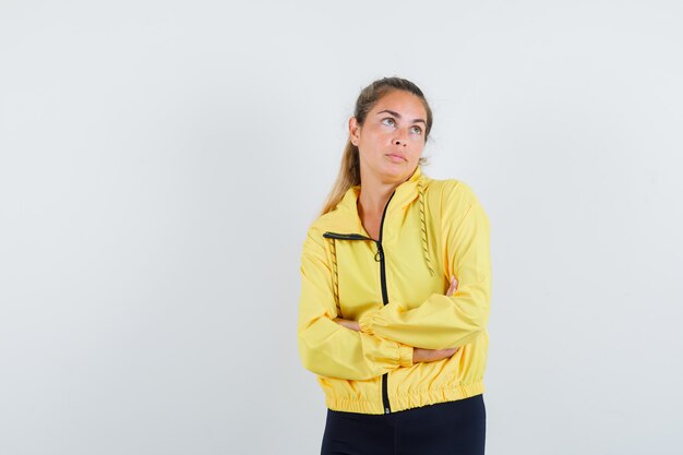 Blonde woman in yellow bomber jacket and black pants standing arms crossed, looking away and looking pensive