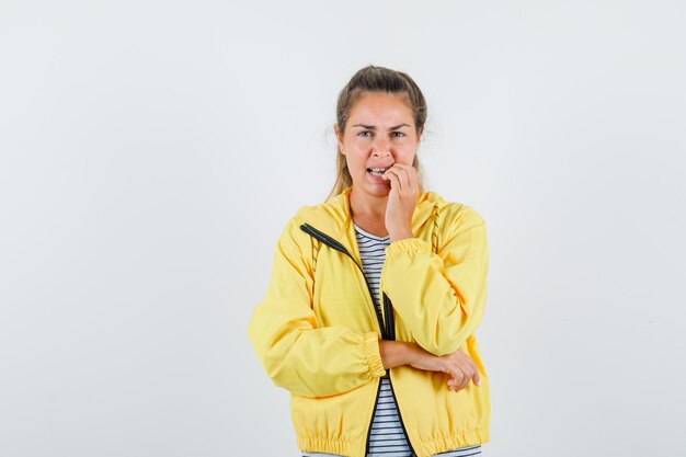 Blonde woman in yellow bomber jacket and striped shirt biting fingers while standing in thinking pose and looking pensive