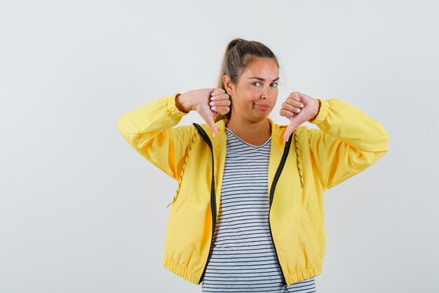 Blonde woman in yellow bomber jacket and striped shirt showing thumbs down with both hands and looking pretty