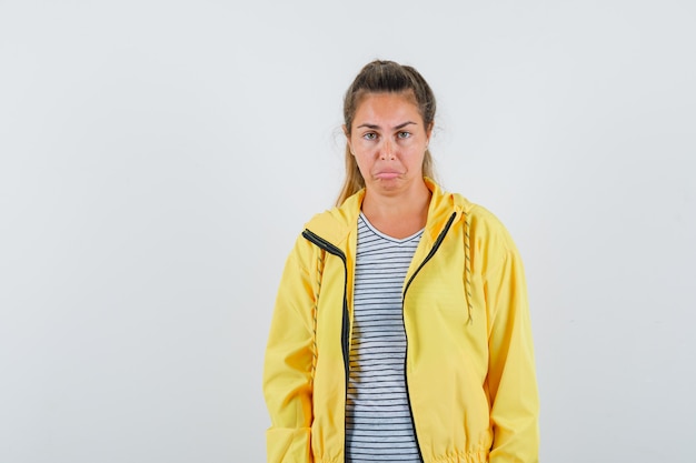 Donna bionda in bomber giallo e camicia a righe in piedi dritta, smorfie e posa alla macchina fotografica e sembra carina