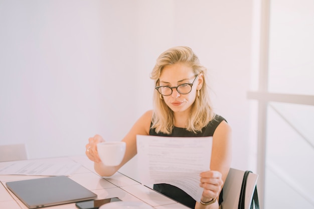 Blonde young businesswoman holding cup of coffee reading the document at workplace