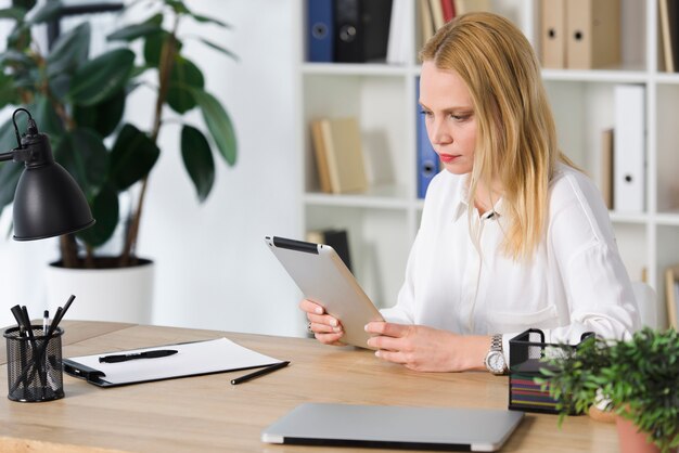 Blonde young businesswoman sitting at workplace looking at digital tablet in the office