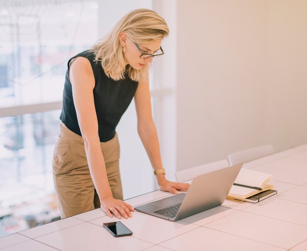 Free photo blonde young businesswoman standing near the desk looking at laptop