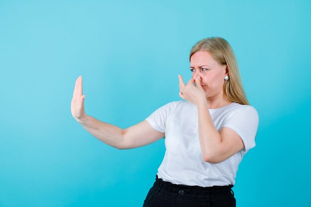 Blonde young girl is holding nose hand showing stop gesture on blue background