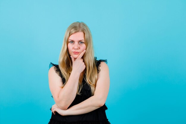 Blonde young girl is looking at camera by putting hand on chin on blue background