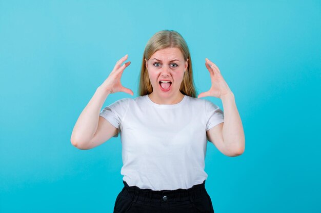 Blonde young girl is screaming by holding hands near head on blue background