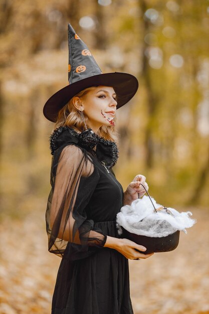 Blonde young girl witch in forest on Halloween. Girl wearing black dress and cone hat. Witch holding a magician stuff.