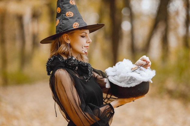 Blonde young girl witch in forest on Halloween. Girl wearing black dress and cone hat. Witch holding a magician stuff.