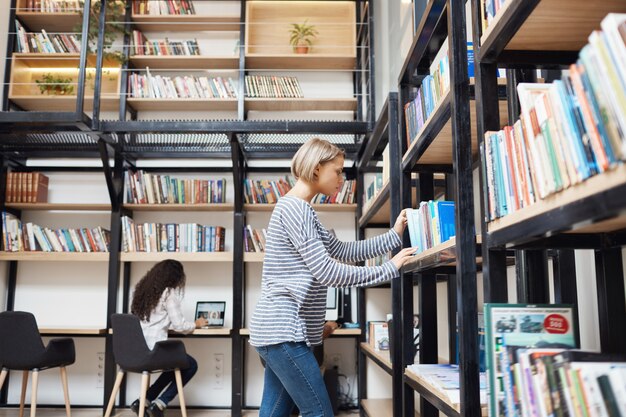 Блондинка молодая красивая женщина в полосатой рубашке и джинсах ищет книгу на полке в библиотеке, готовясь к экзаменам в университете
