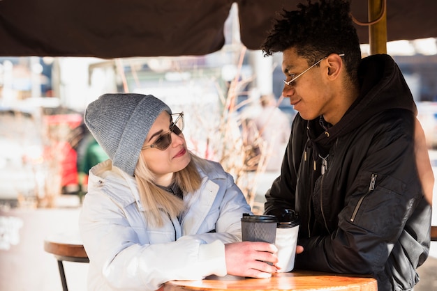 Blonde young interracial young couple holding disposable coffee cup sitting in caf�