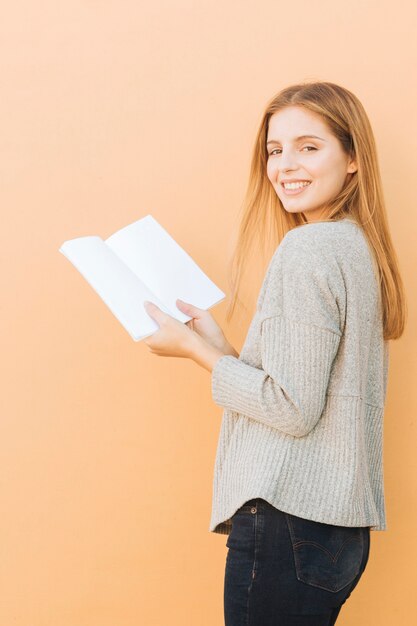 Белокурая молодая женщина держа книгу в руке смотря камеру против предпосылки персика