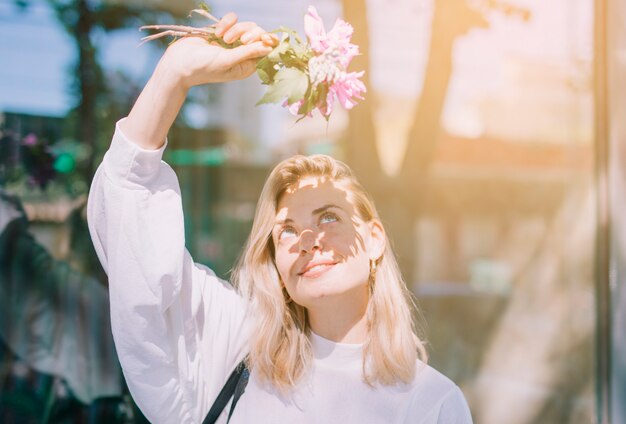 Blonde young woman holding flowers in hand shielding her eyes from the sunlight