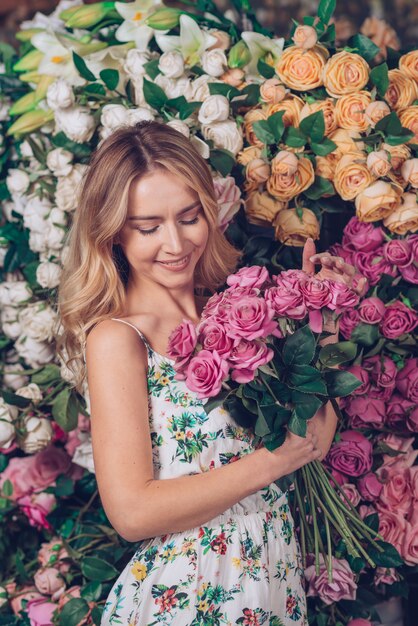 Blonde young woman holding pink roses in hand standing in front of flower backdrop