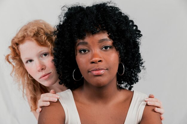 Blonde young woman peeking from an african female friend against grey backdrop