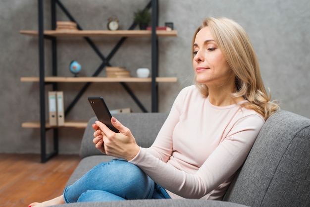 Белокурая молодая женщина сидя на софе используя умный телефон дома